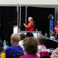 Deputy Katie Sherrod of North Texas speaks to a resolution that reunited her diocese with the Diocese of Texas. General Convention volunteer Becky Walker, in red vest, looks on.